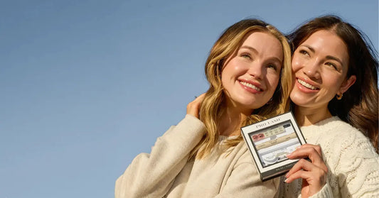 Lash extension kit held by two smiling women outdoors under a clear blue sky
