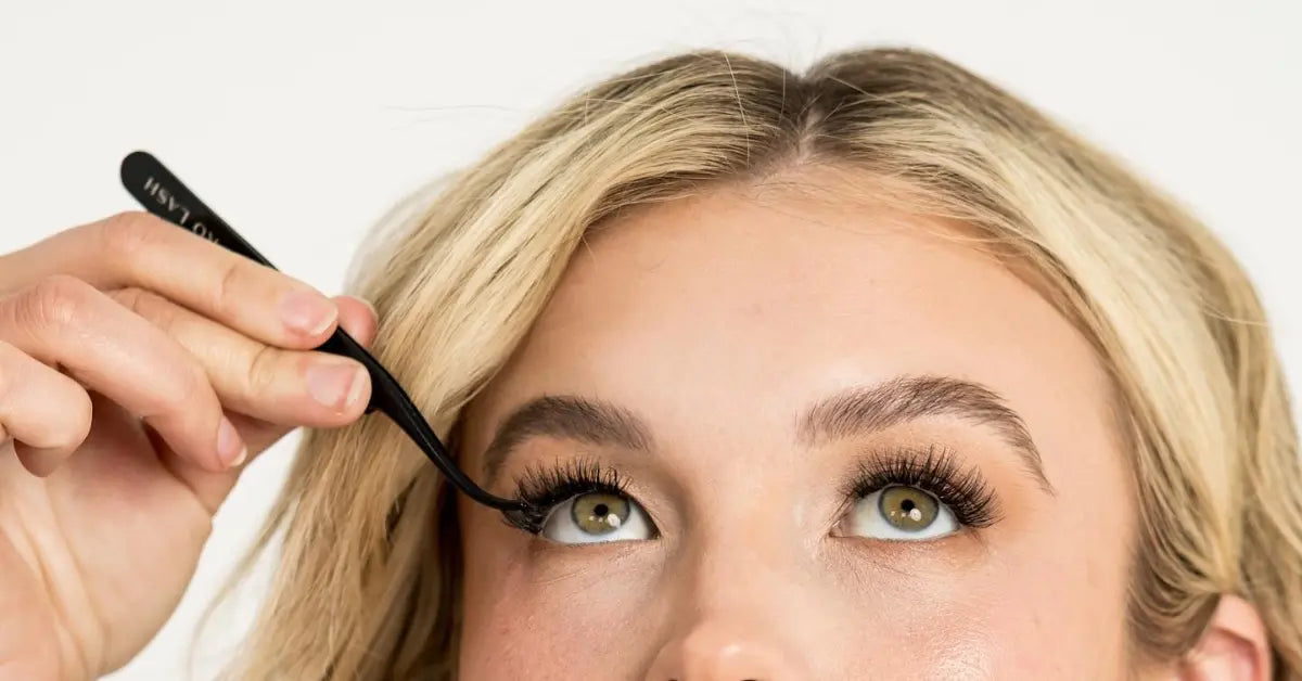 Volume lashes being applied with black tweezers to upper eyelid of a woman with blonde hair.